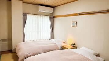 A hotel room with two beds, a bedside table with a lamp, an air conditioning unit, and a window with curtains.