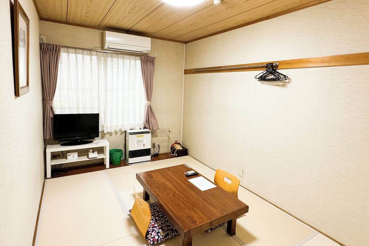 A room with a wooden ceiling, a television on a stand, a table with chairs, and a window with curtains.