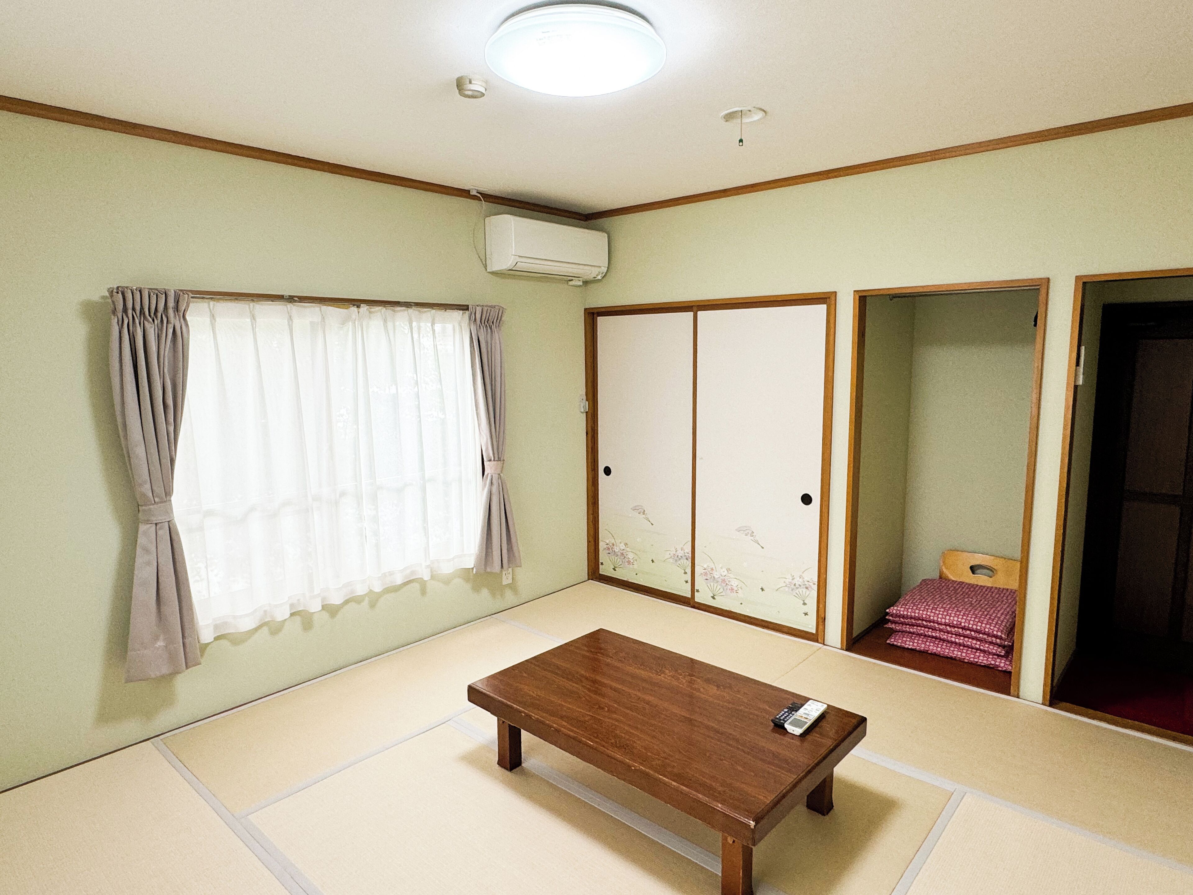 Una habitación con suelo de tatami, una mesa de madera, un ventanal con cortinas, un puertas corredera con un diseño floral y un equipo de aire acondicionado.