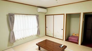 Ein Zimmer mit Tatami-Böden, einem Holztisch, einem Vorhangfenster, einer Holztür mit floralem Dekor und einer Klimaanlage.