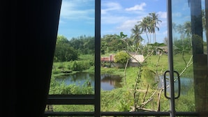 A view through a window with a curtain, featuring a lush green landscape, a body of water, and palm trees.