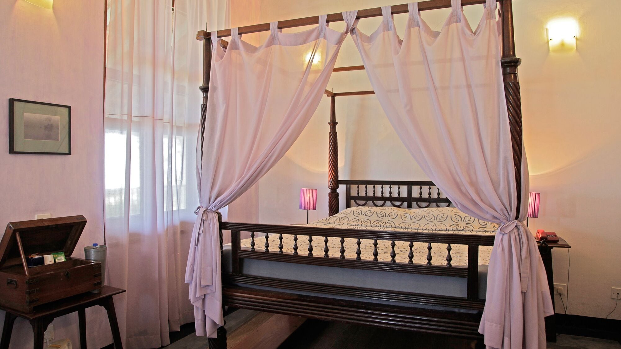 A four-poster bed with a canopy, a bedside table, a lamp, and a framed picture on the wall.