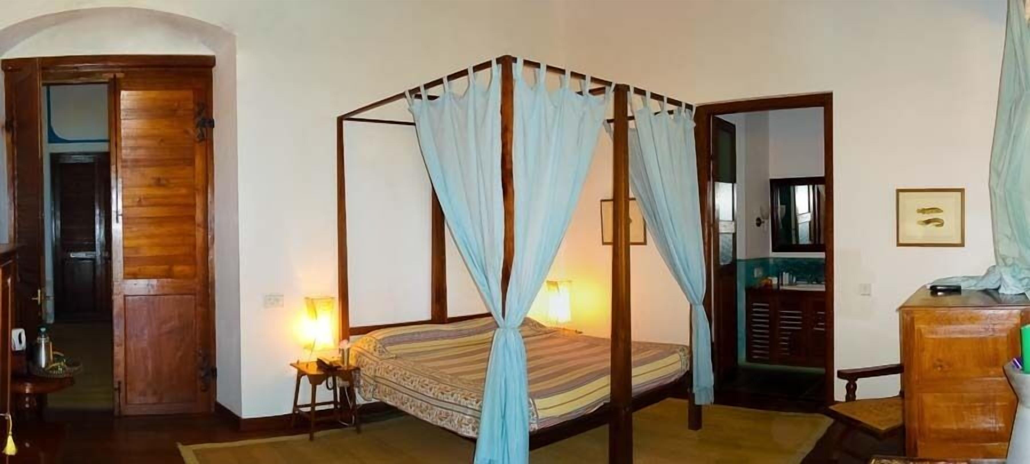 A four-poster bed with a canopy, a wooden wardrobe, a mirror, and a small table with a lamp.
