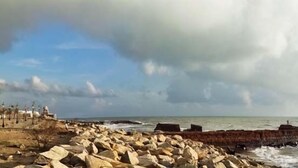 Una zona costanera amb un tram d'embarcador rocós, un edifici amb teulada vermella i un cel amb alguns núvols.