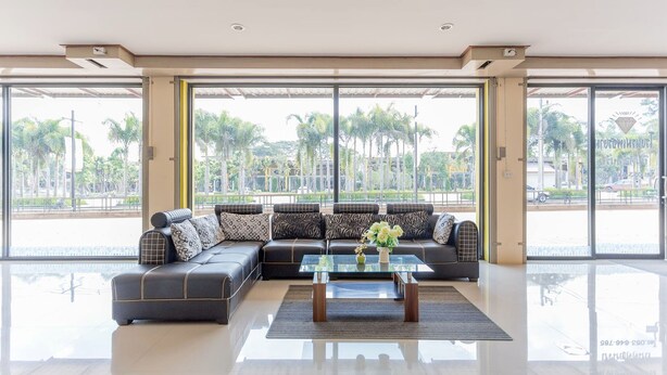 A modern living room with a sectional sofa, a glass coffee table, and large windows offering a view of palm trees and a road. - Phet Ngam Hotel (Mae Sai)