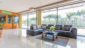 A spacious lobby with a black leather sofa, a glass coffee table, and large windows offering a view of palm trees and a parking area. - Phet Ngam Hotel (Mae Sai)