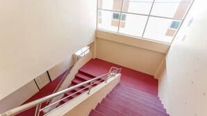 A staircase with a red carpet and metal handrails, leading to a landing with a window overlooking a building. - Phet Ngam Hotel (Mae Sai)