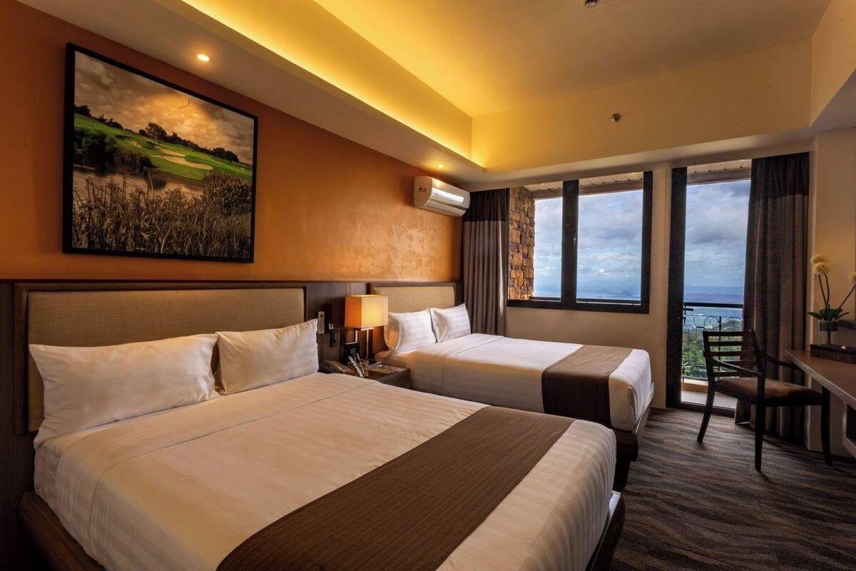 A hotel room with two beds, a desk, a chair, a painting, and a view of the outdoors.