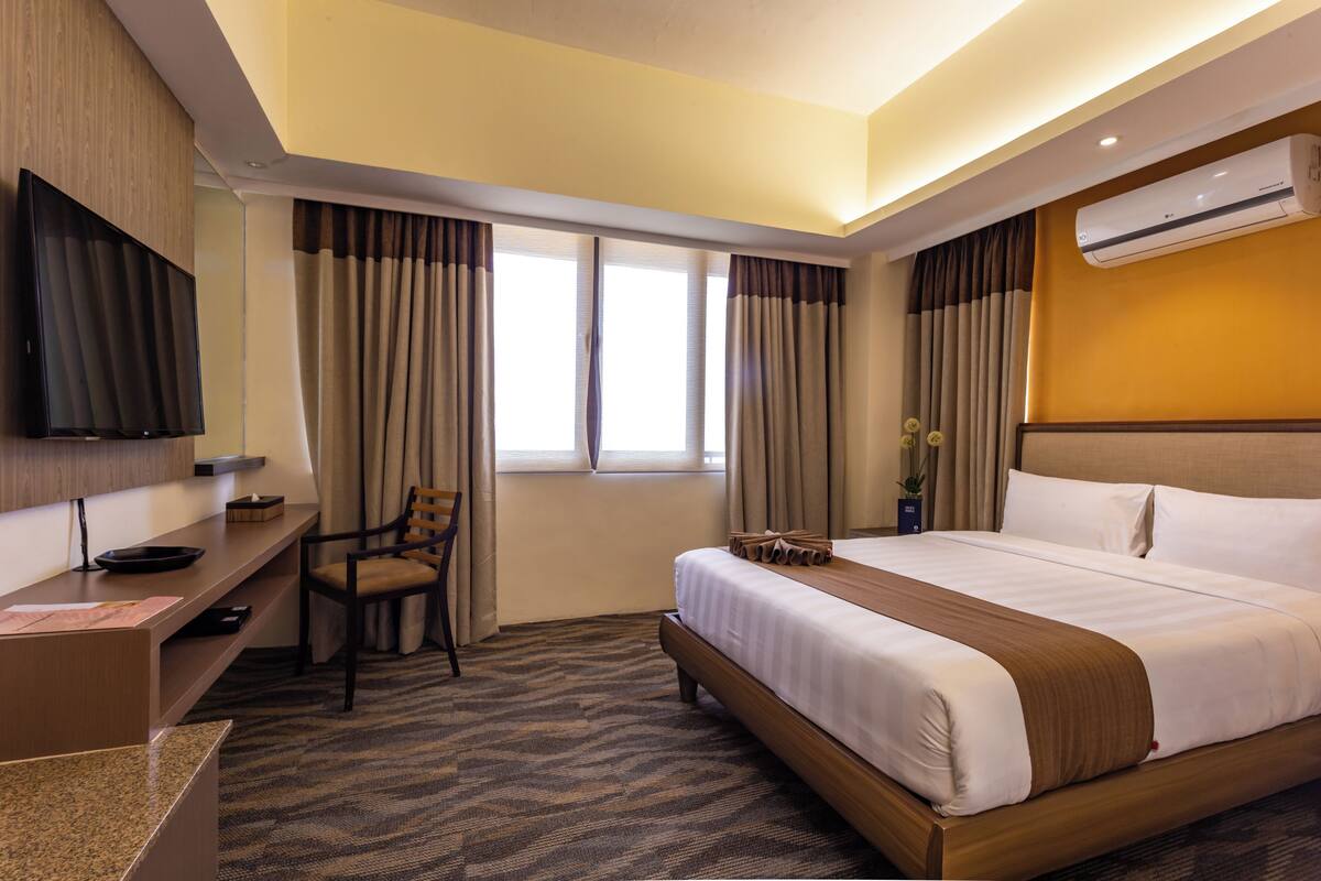 A hotel room with a bed, desk, chair, TV, and an air conditioning unit.