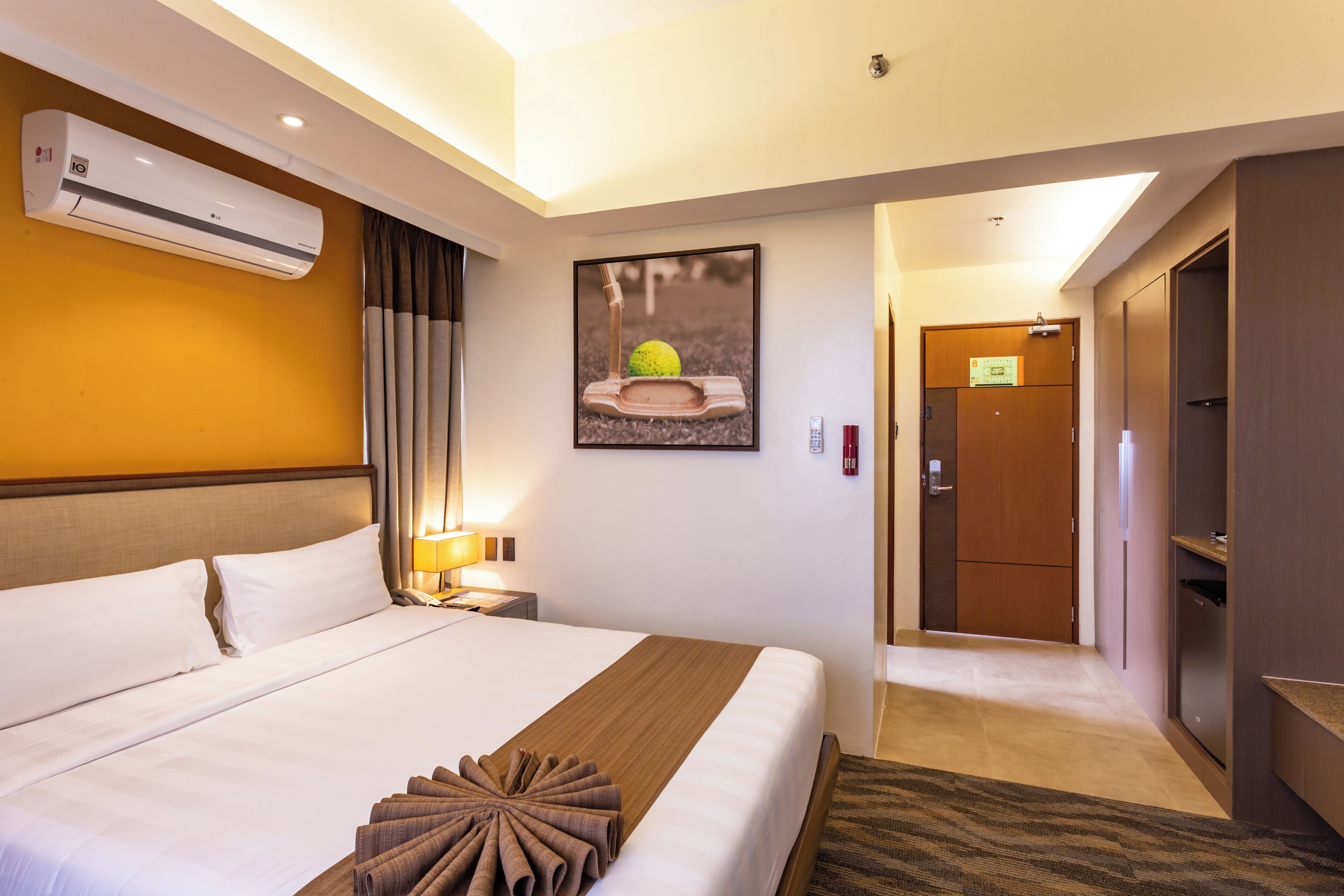 A hotel room with a large bed, a wall-mounted air conditioner, a framed picture, a door, and a small table with a lamp.