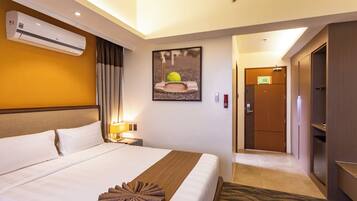 A hotel room with a large bed, a wall-mounted air conditioner, a framed picture, a door, and a small table with a lamp.