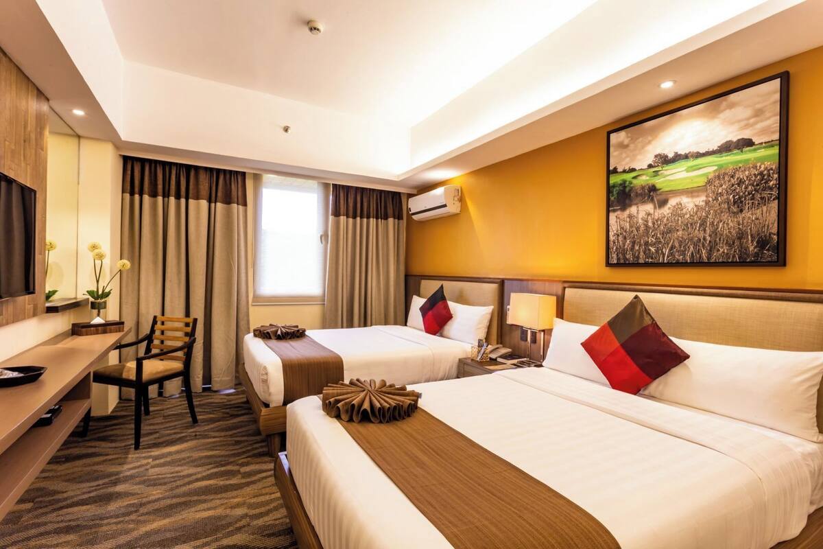 A hotel room with two beds, a desk, a chair, a large window with curtains, an air conditioning unit, and a painting on the wall.