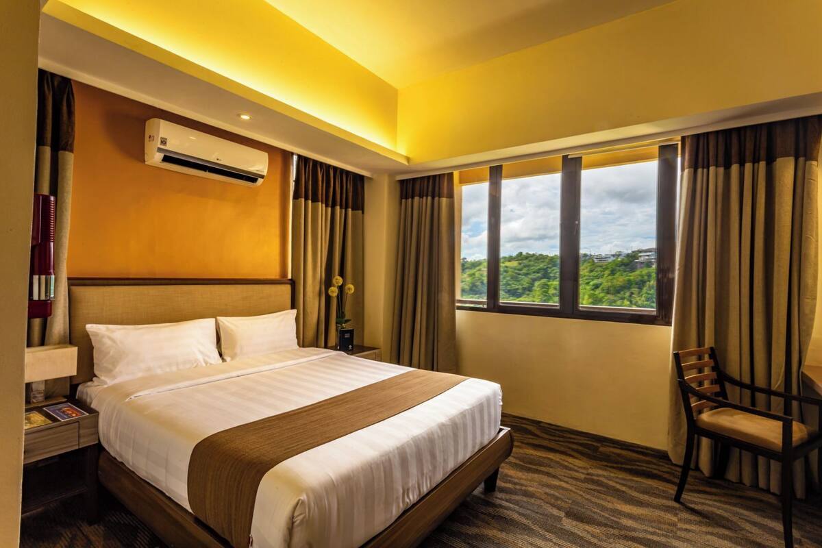A hotel room with a large bed, a nightstand with a lamp, an air conditioner, a window with curtains, and a view of greenery outside.
