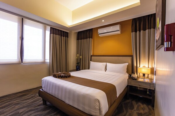A hotel room with a large bed, a bedside table with a lamp, an air conditioning unit, and a window with curtains.