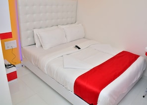 A neatly made bed with white bedding and a red accent, a remote control on the bed, and a power outlet on the wall. - Hotel Blue Sea Residency (Mumbai)