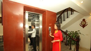 Interior de un ascensor con una persona parada afuera, una escalera con barandilla de madera y una planta en maceta.