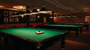 A pool hall with multiple green pool tables, red cushioned seating, and pendant lighting.
