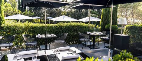 An outdoor dining area with tables and chairs, surrounded by greenery and umbrellas.