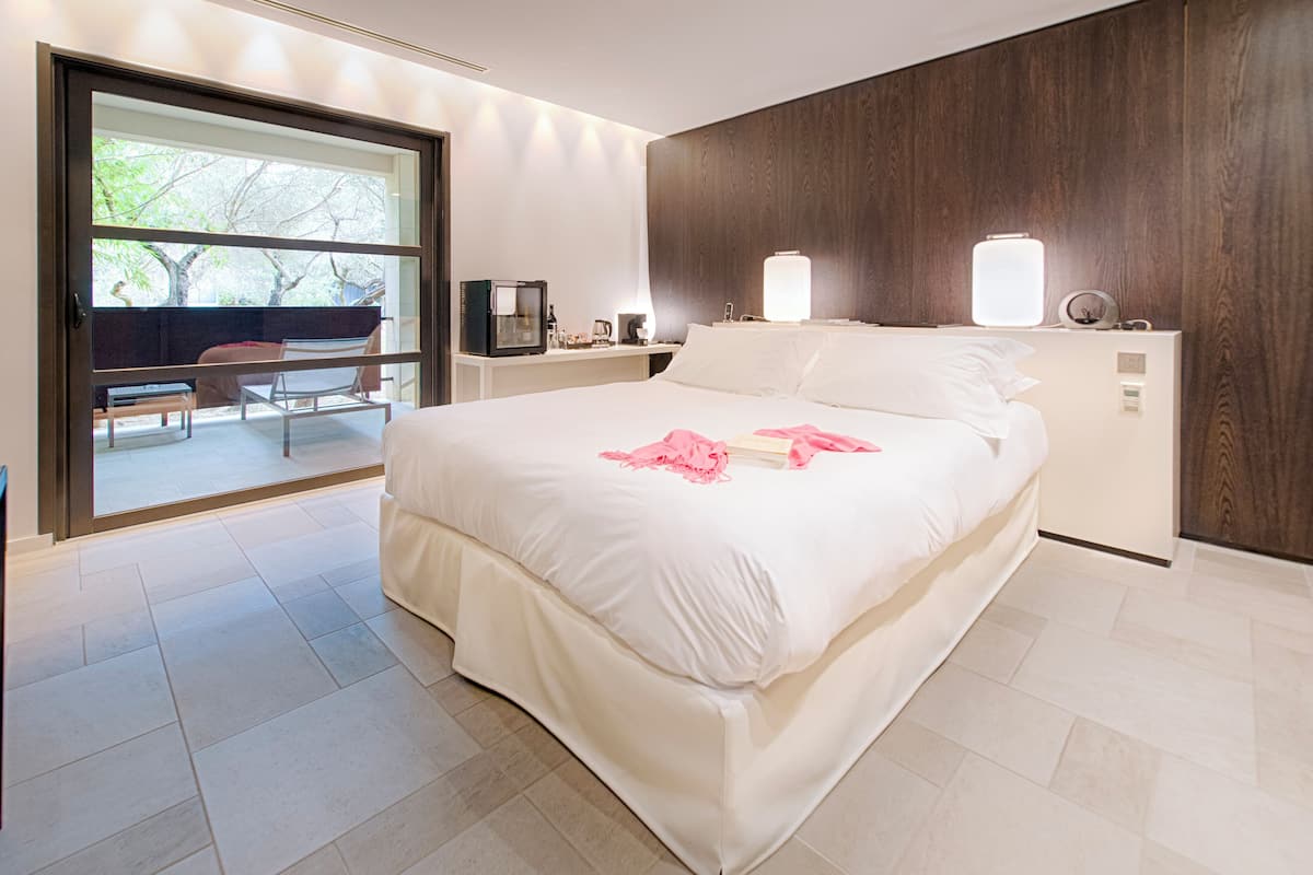 A modern hotel room with a large bed, a wooden headboard, a bedside table with a lamp, and a view through a glass door to an outdoor seating area.