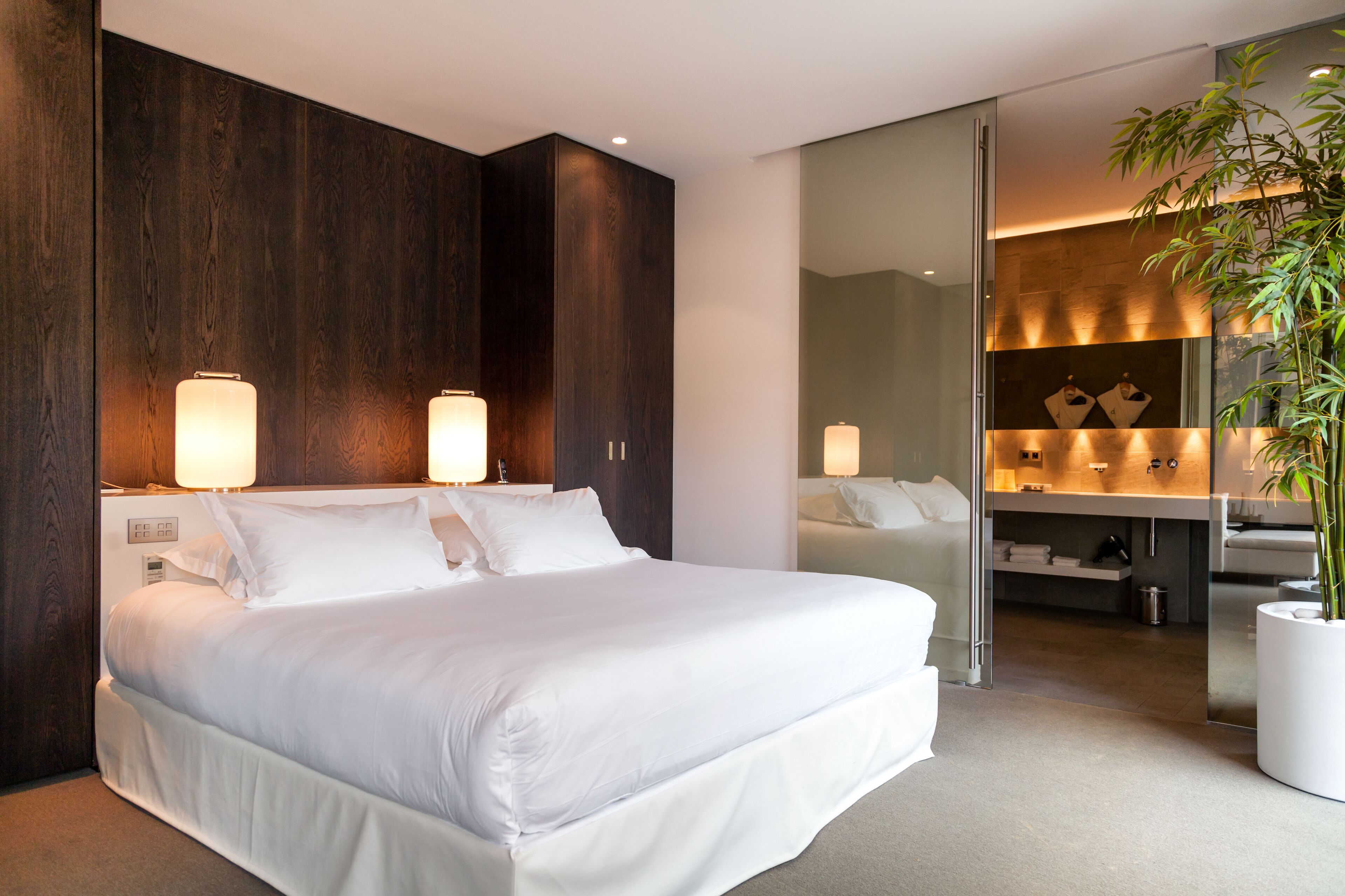 Un dormitorio moderno con una cama grande, cabecera de madera, lámparas de noche, un espejo, una planta y un vestidor.