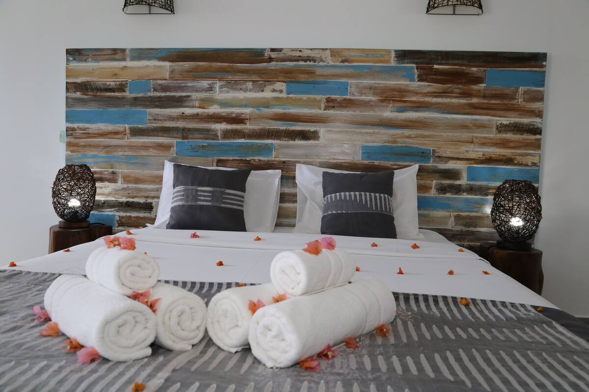 A neatly made bed with rolled white towels and pink flowers, a patterned bedspread, and a rustic wooden headboard.