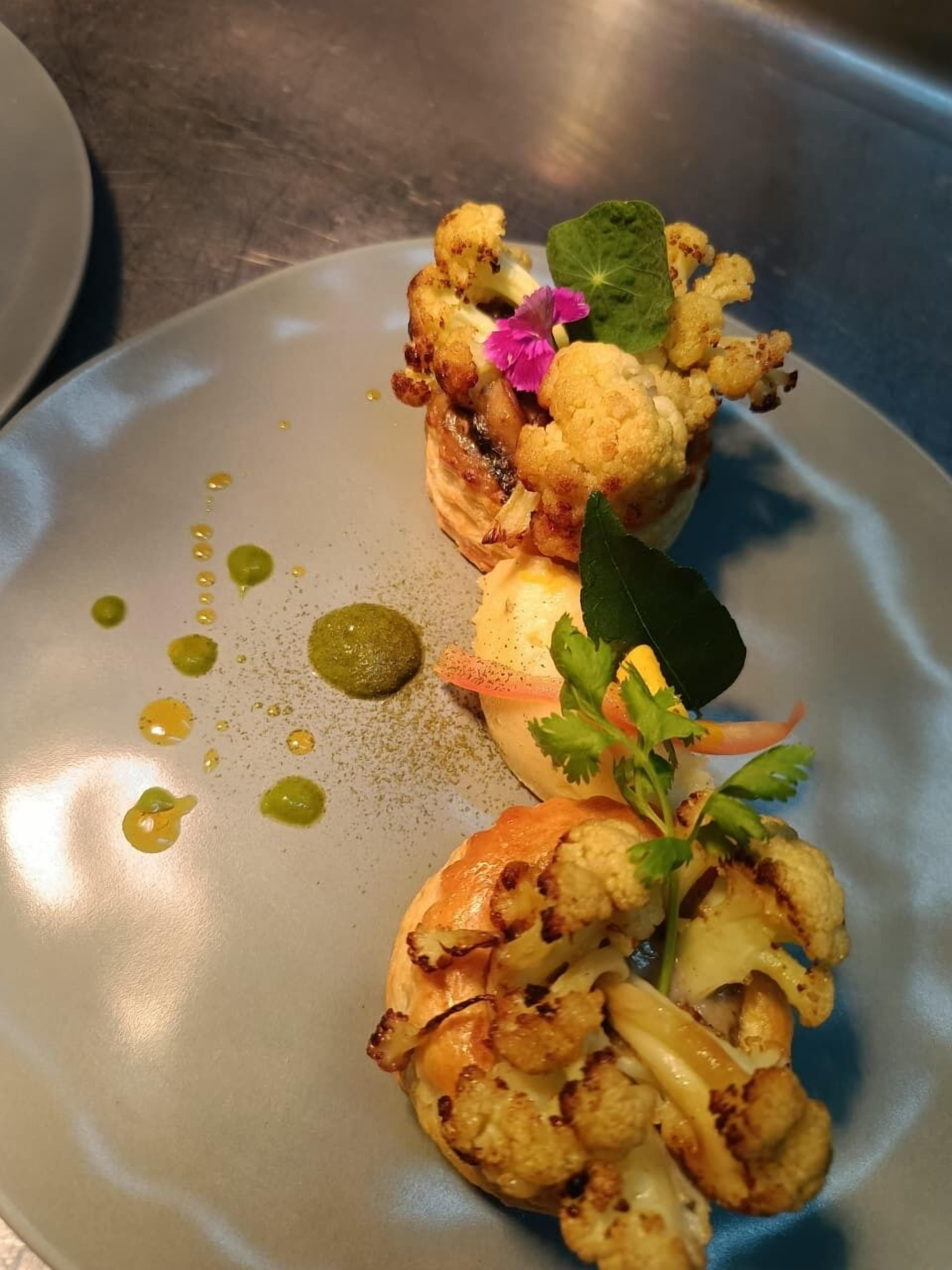Un plato con alimentos, incluyendo coliflor y posiblemente pollo, adornado con hierbas y una flor.