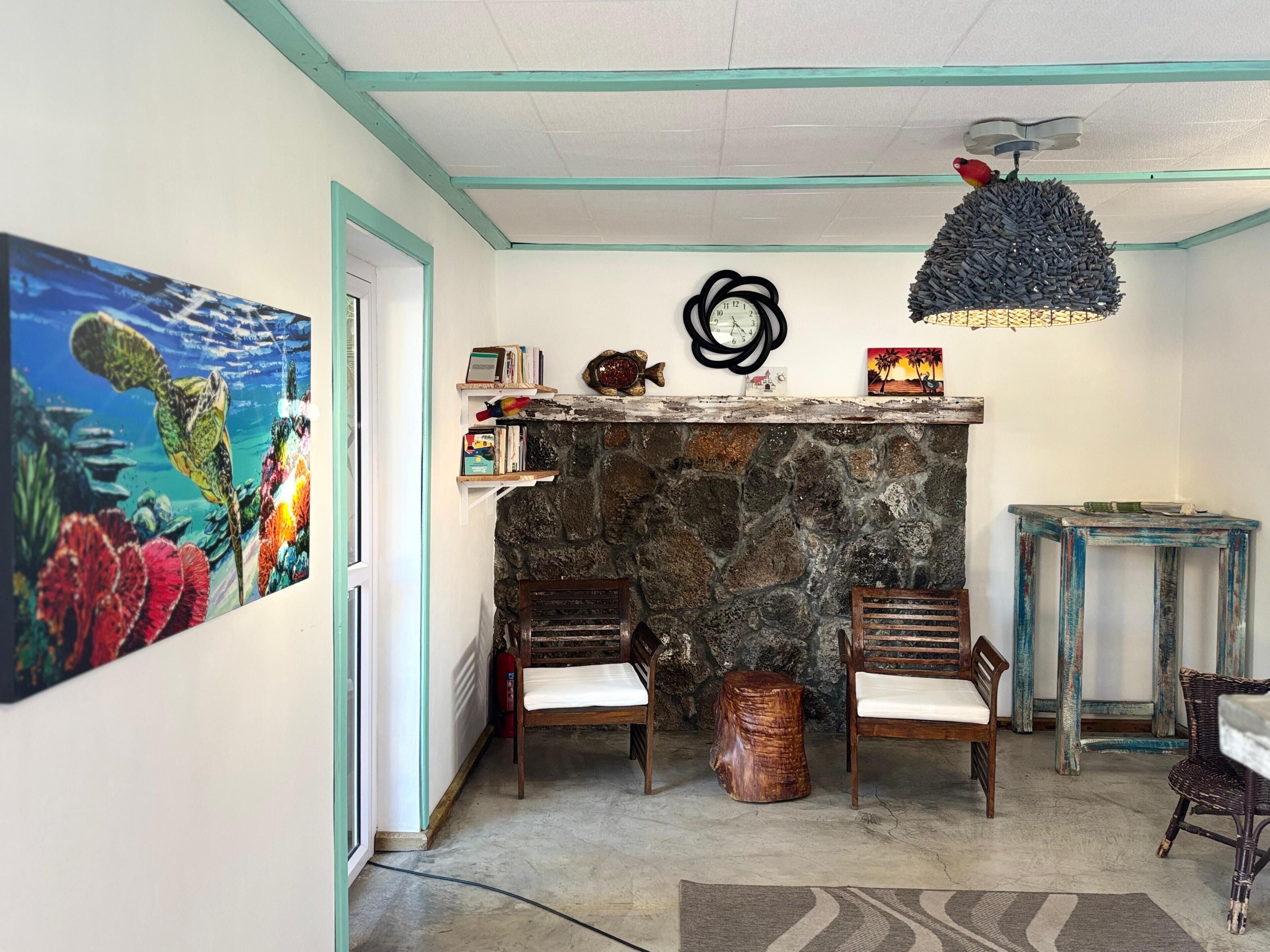 A room with a stone fireplace, two wooden chairs, a small round wooden stool, a wall clock, and a ceiling fan.
