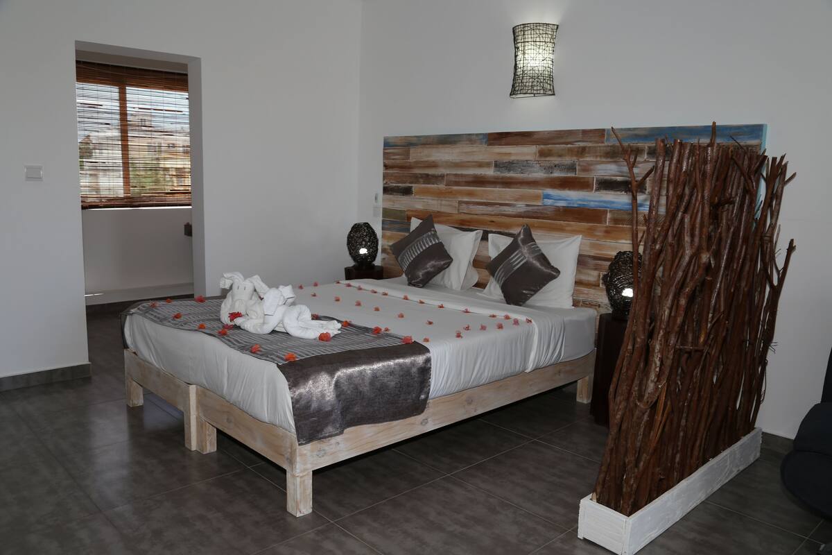 A bedroom with a wooden bed, white bedding, a decorative wall, and a window with blinds.
