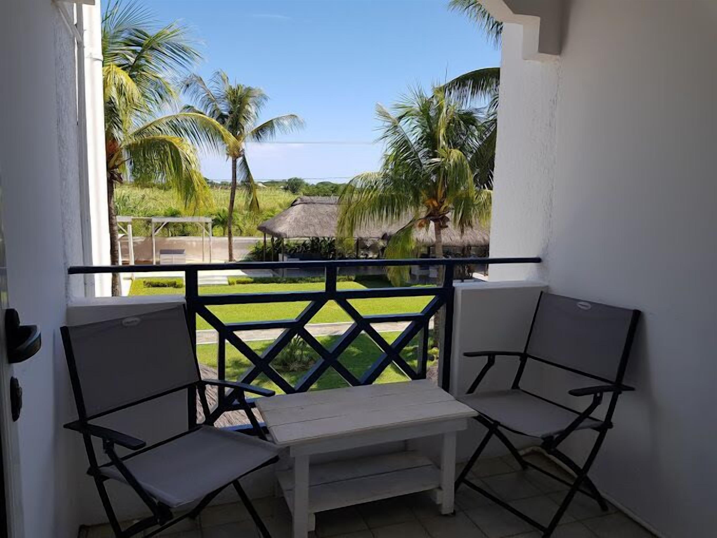 A balcony with two white chairs and a small table, overlooking a grassy area with palm trees and a thatched-roof structure.