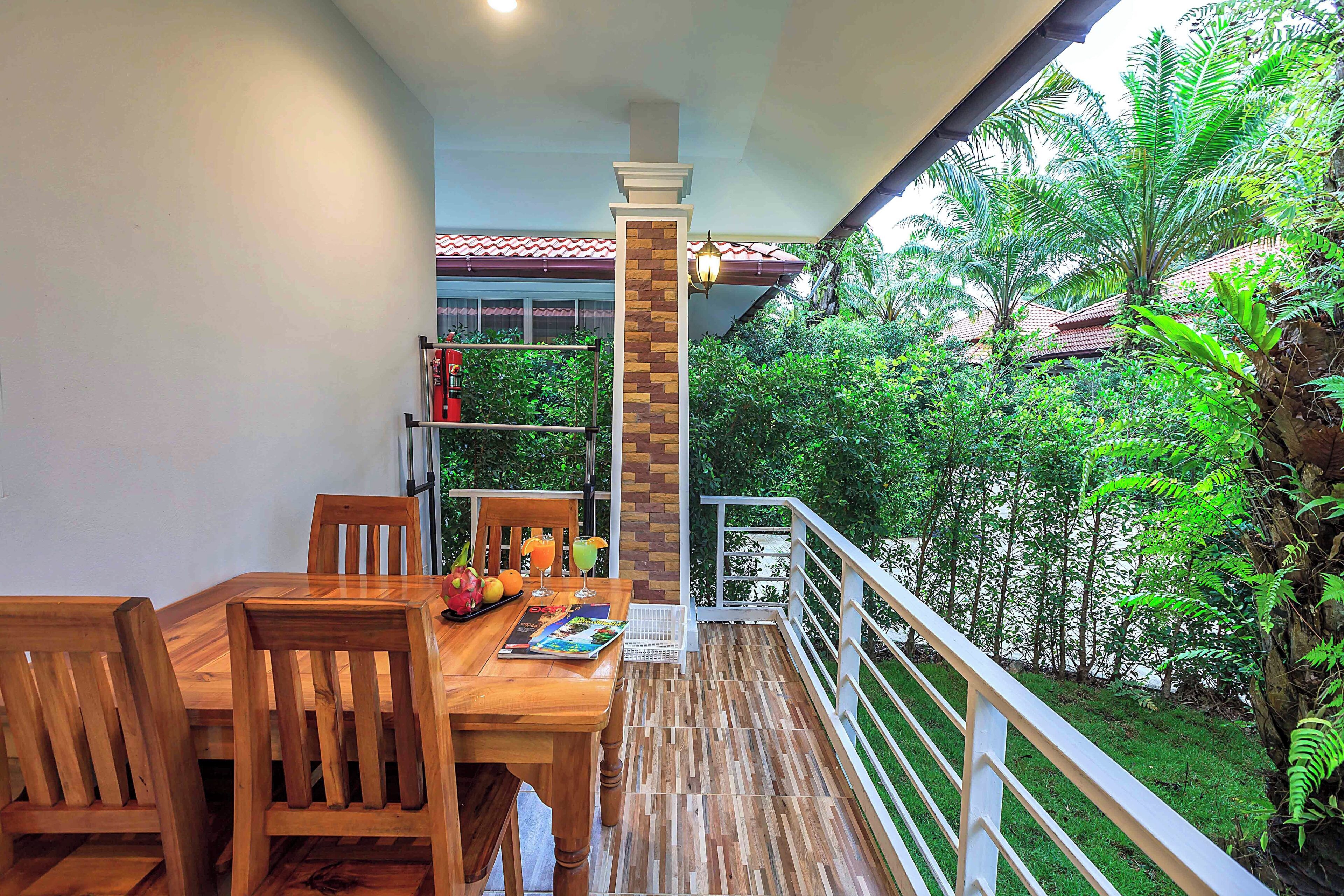 A wooden table with chairs and a magazine, a small shelf with books, and a balcony with a railing overlooking a garden.