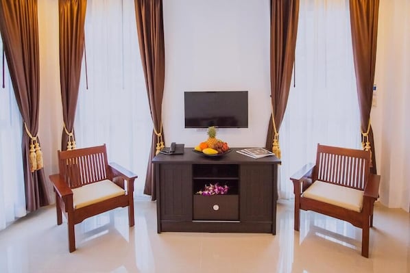Een kamer met een flatscreen televisie aan de muur, een houten bureau met een schaal fruit en een telefoon, twee houten stoelen met kussens, en een klein plankje met decoratieve objecten.