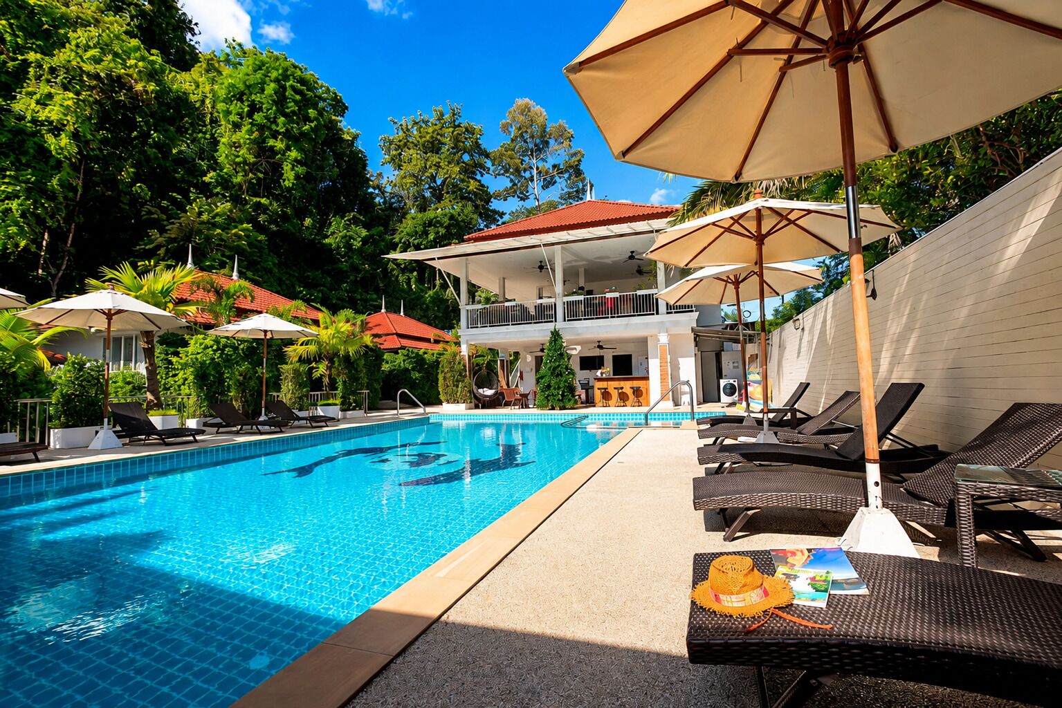 A swimming pool with lounge chairs and umbrellas, surrounded by greenery and a building with a red roof.