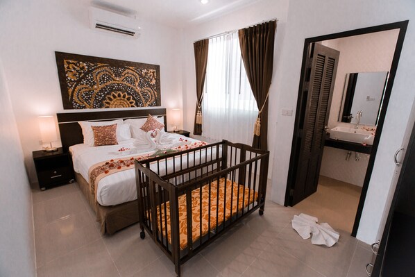 Hotelkamer met een bed, ledikant, nachtkastje, een ingebouwde airconditioning, een badkamer met wastafel en spiegel, en een deur die leidt naar een andere kamer.