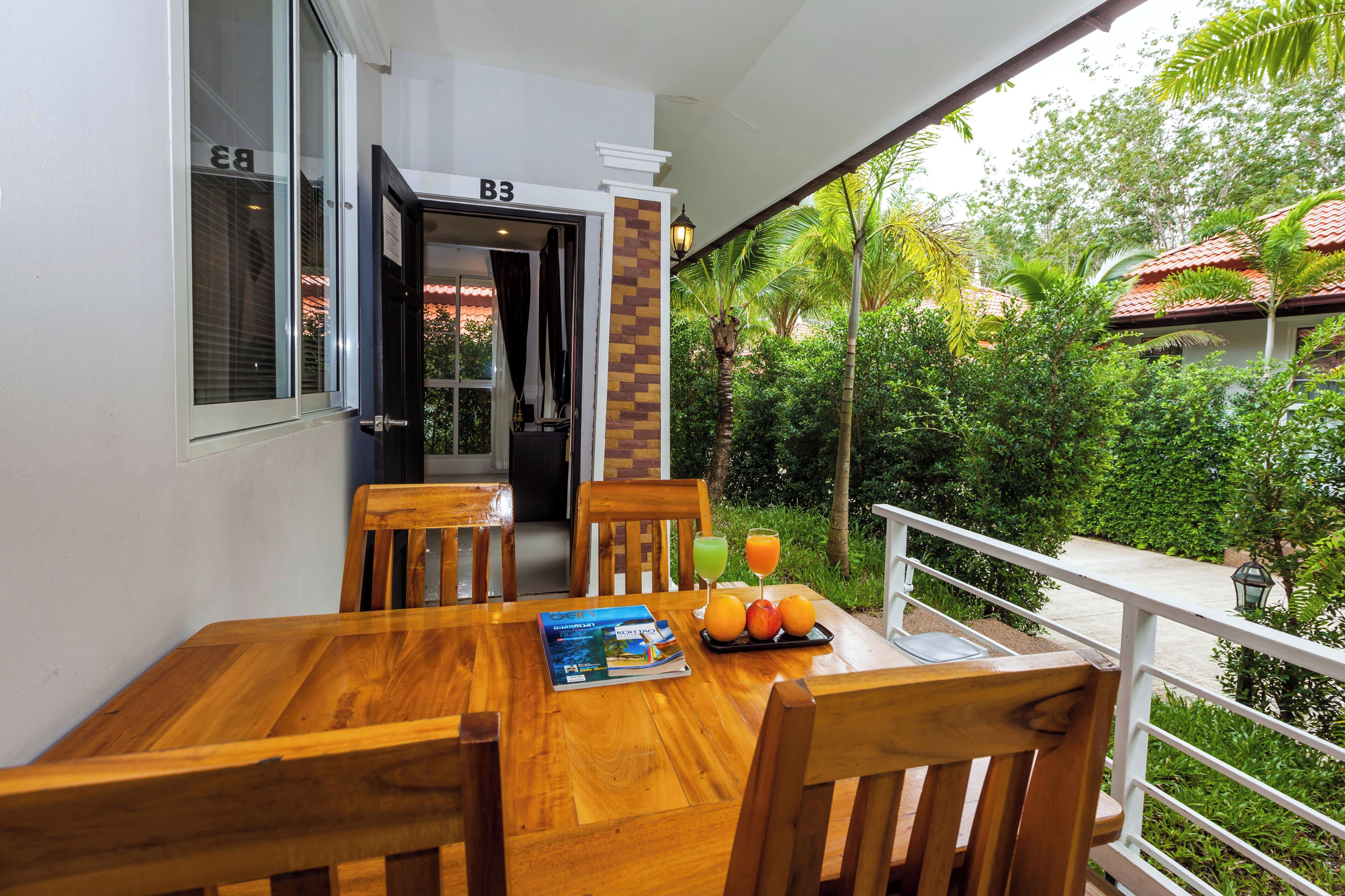 Una mesa de comedor de madera con sillas, una revista y fruta en un balcón con vistas a zonas verdes y un edificio con techo rojo.