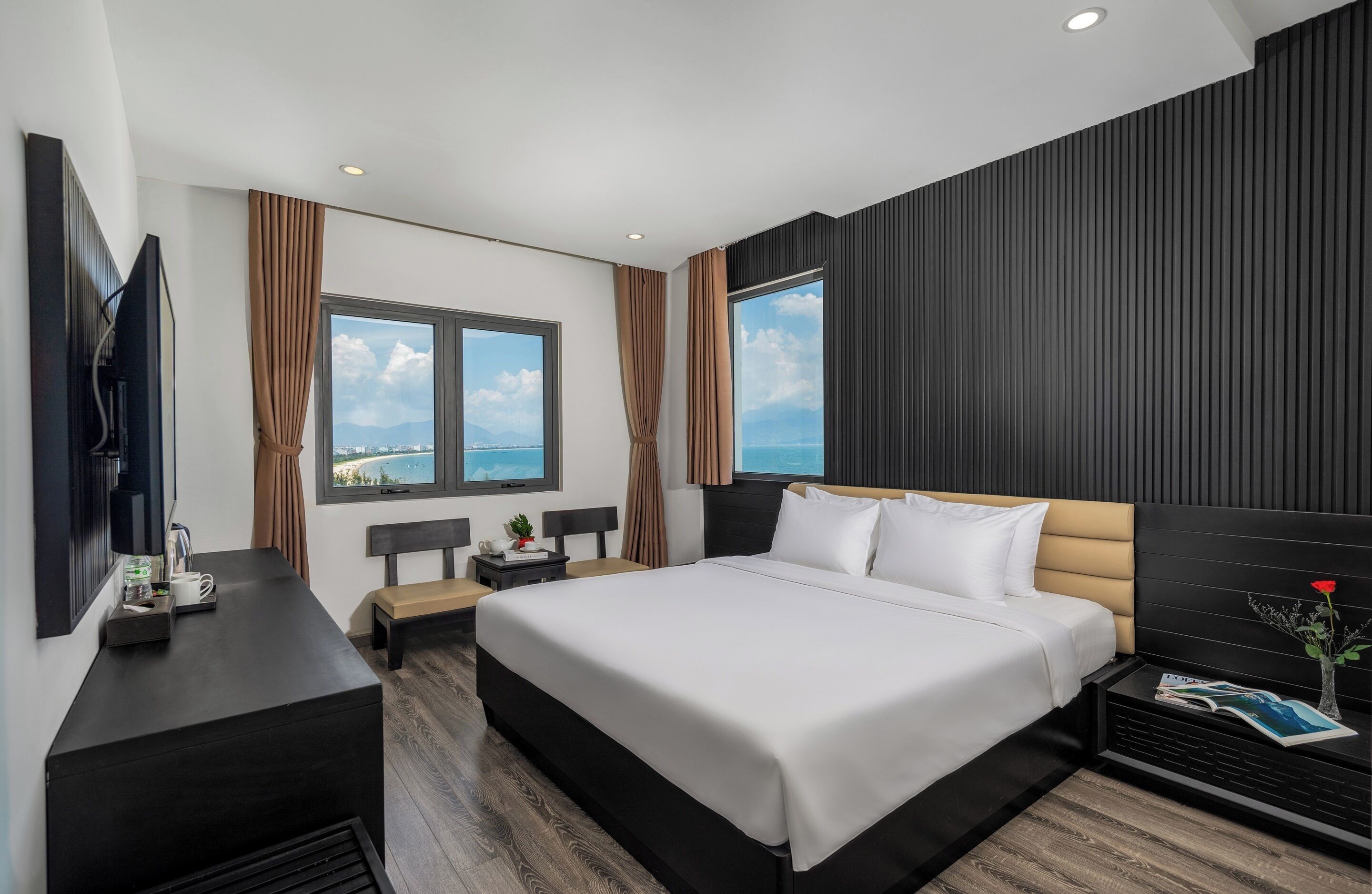 A modern hotel room with a large bed, a flat-screen TV, a bedside table with a lamp, and a view of the sea through large windows.