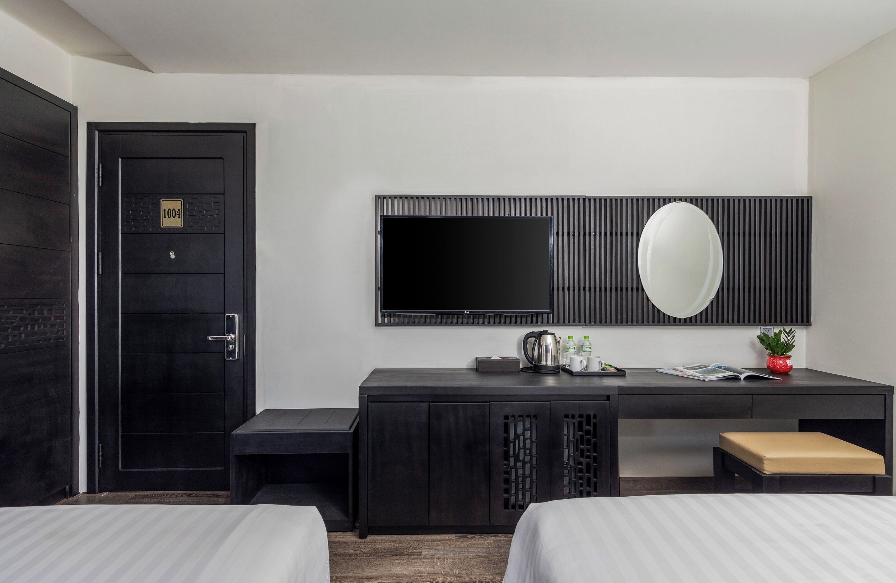 Habitación de hotel con una puerta de madera oscura, un escritorio negro con televisor de pantalla plana, un espejo y una planta pequeña. Hay dos camas con ropa blanca.
