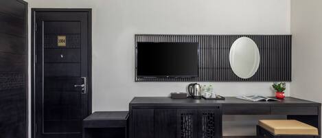 Habitación de hotel con una puerta de madera oscura, un escritorio negro con televisor de pantalla plana, un espejo y una planta pequeña. Hay dos camas con ropa blanca.