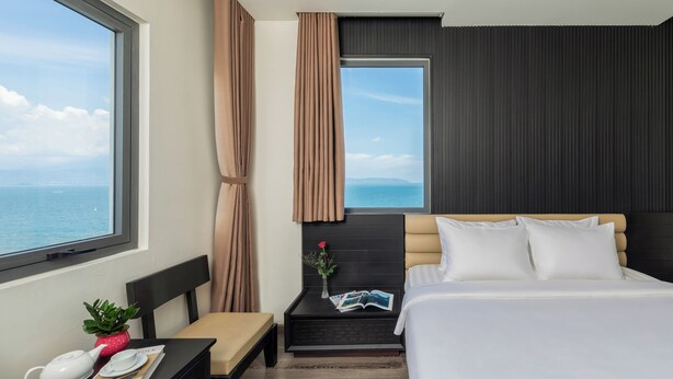 A hotel room with a bed, a bedside table with a book and a teapot, a small table with a plant, and a window with a view of the sea. - Ocean Star Da Nang Bay Hotel (Da Nang)