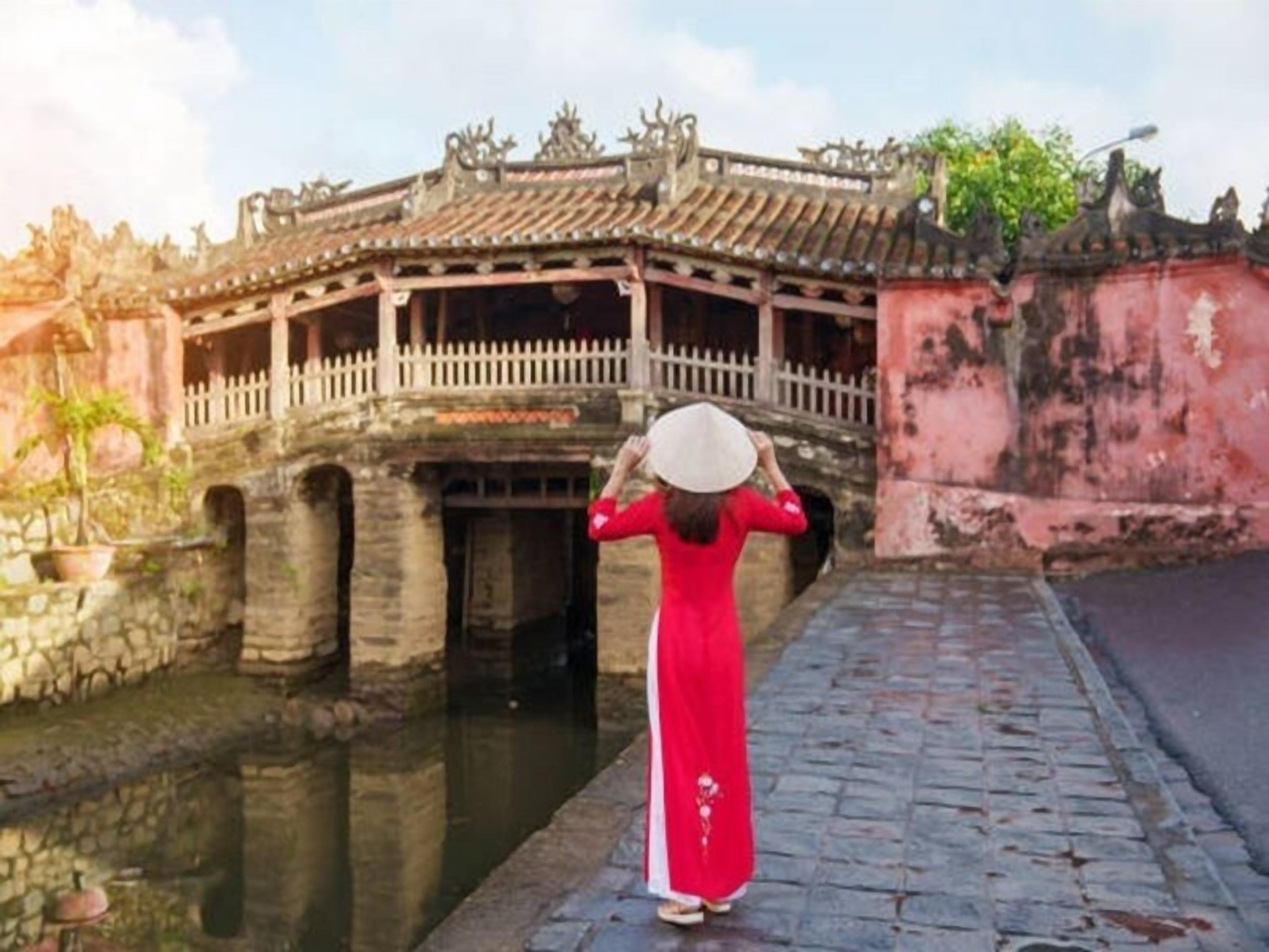 A woman in a red dress and white hat standing in front of a traditional building with a tiled roof and a white fence.