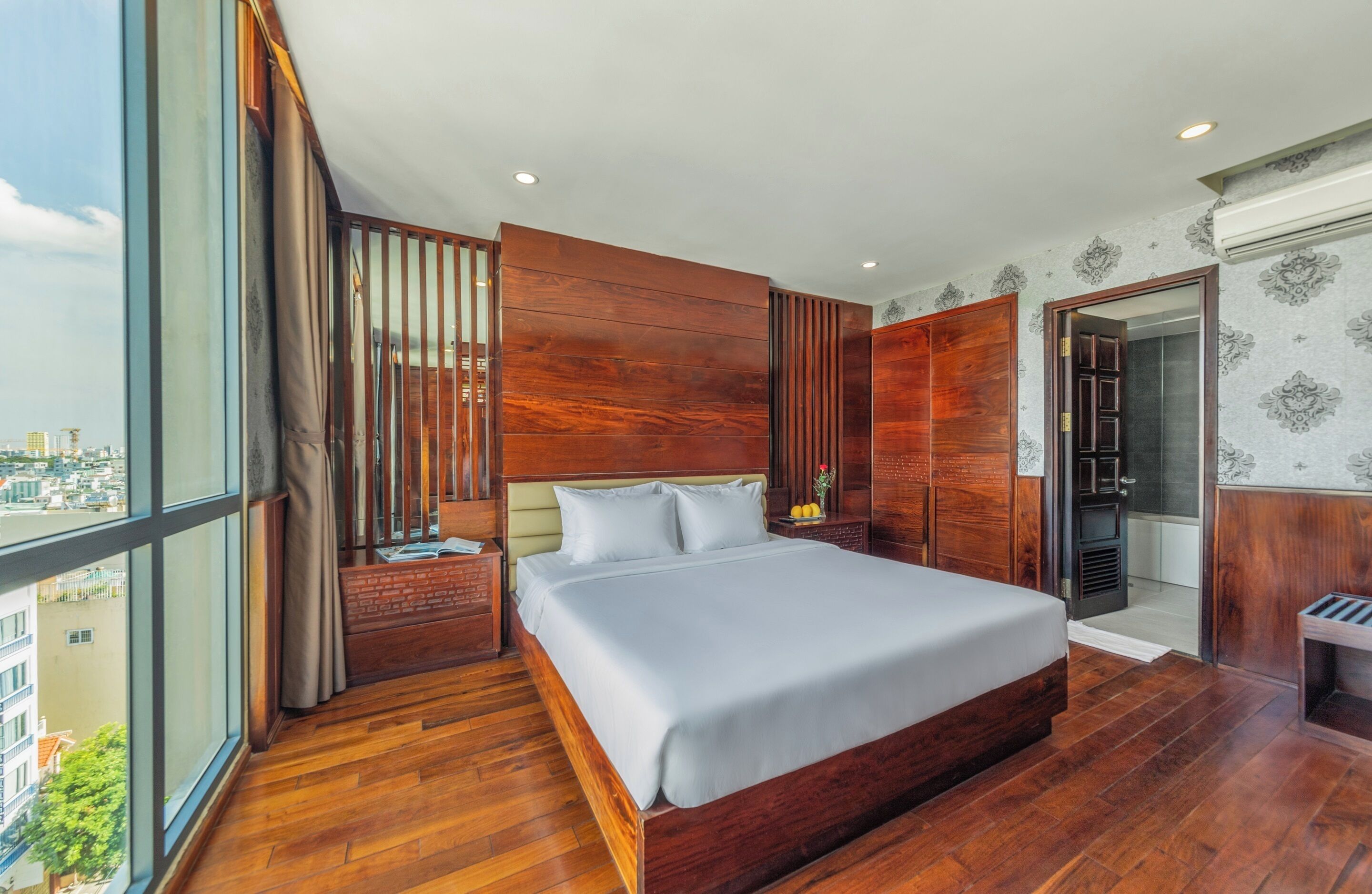 A modern bedroom with a large bed, wooden paneling, a nightstand, and a view of the city.