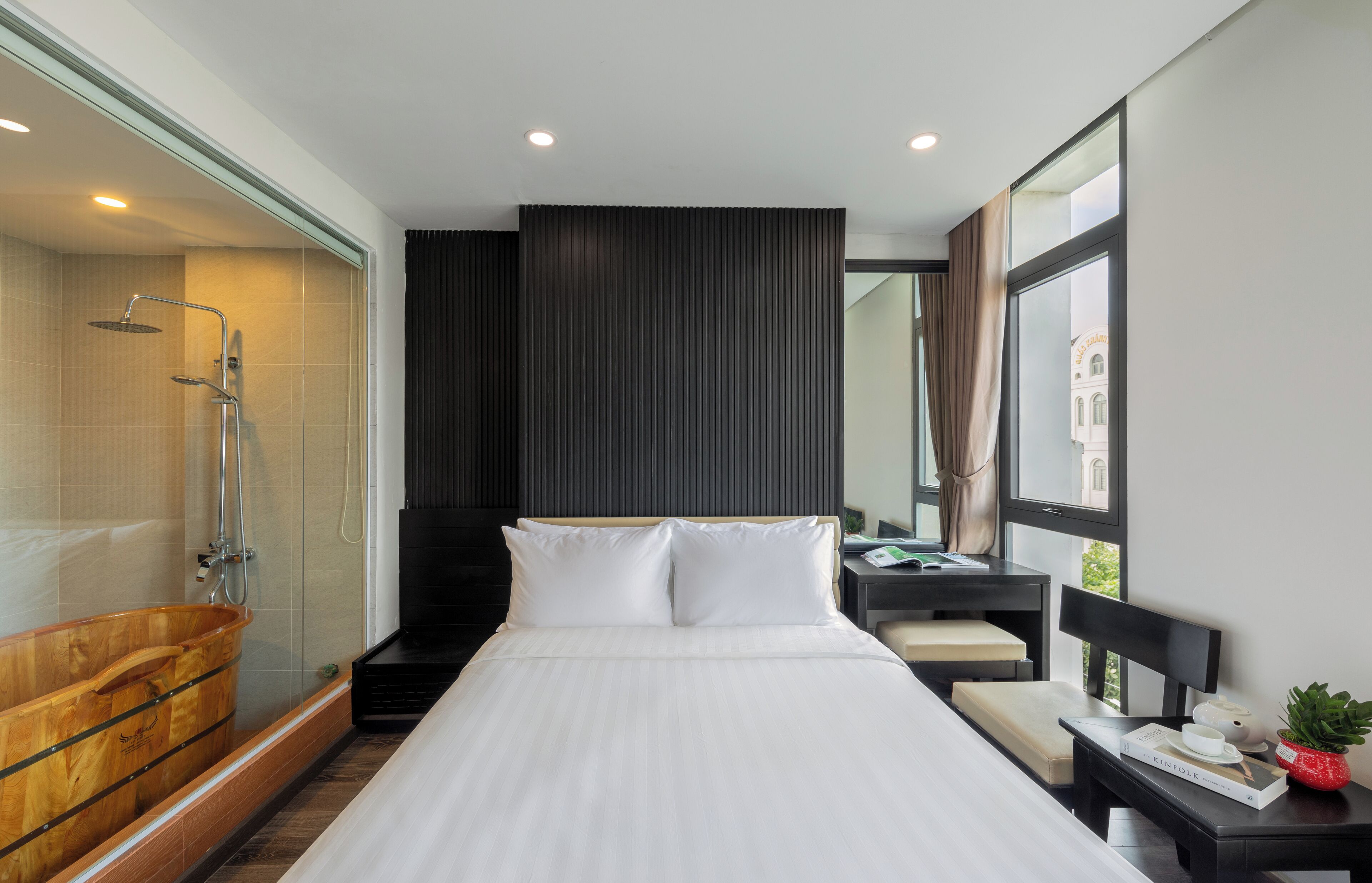 A modern hotel room with a large bed, a wooden bathtub, a glass-enclosed shower, and a view of a building through the window.