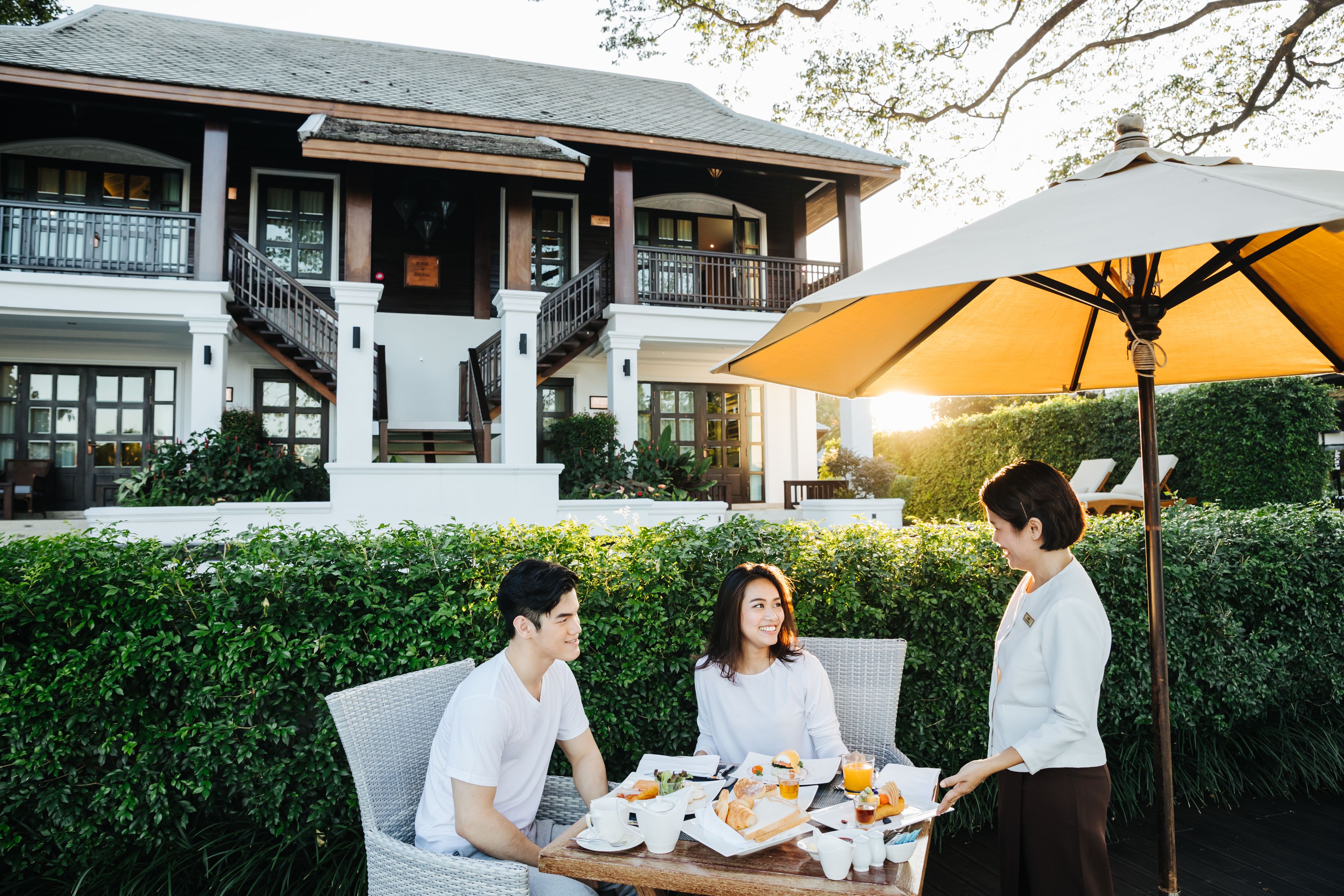 A dining area with a table set for two, a waiter, and a large umbrella providing shade.