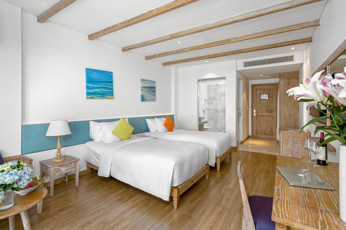 A hotel room with two beds, a wooden floor, a desk with a laptop, a vase with flowers, and artwork on the wall.