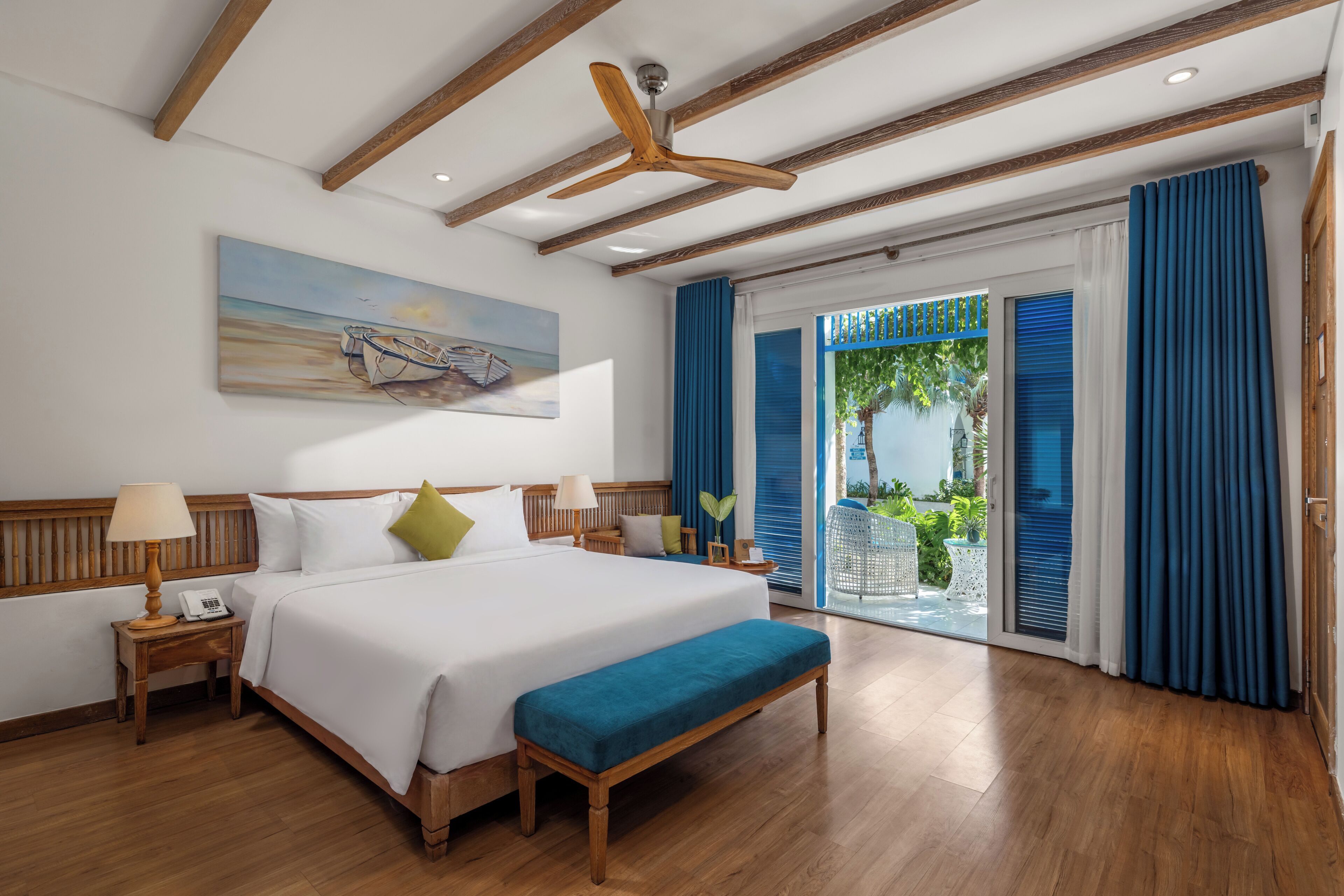 A hotel room with a large bed, wooden headboard, a ceiling fan, a painting of boats, a balcony with blue curtains, and a view of greenery.