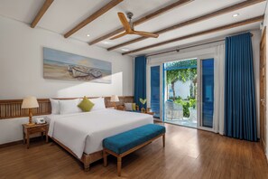 Quarto de hotel com cama grande, cabeceira de madeira, ventoinha no teto, pintura de barcos, varanda com cortinas azuis e vista para o verde.