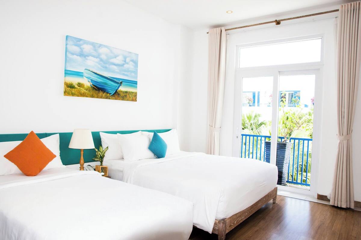 A hotel room with two beds, a painting of a beach scene, a balcony with blue railings, and a view of greenery outside.