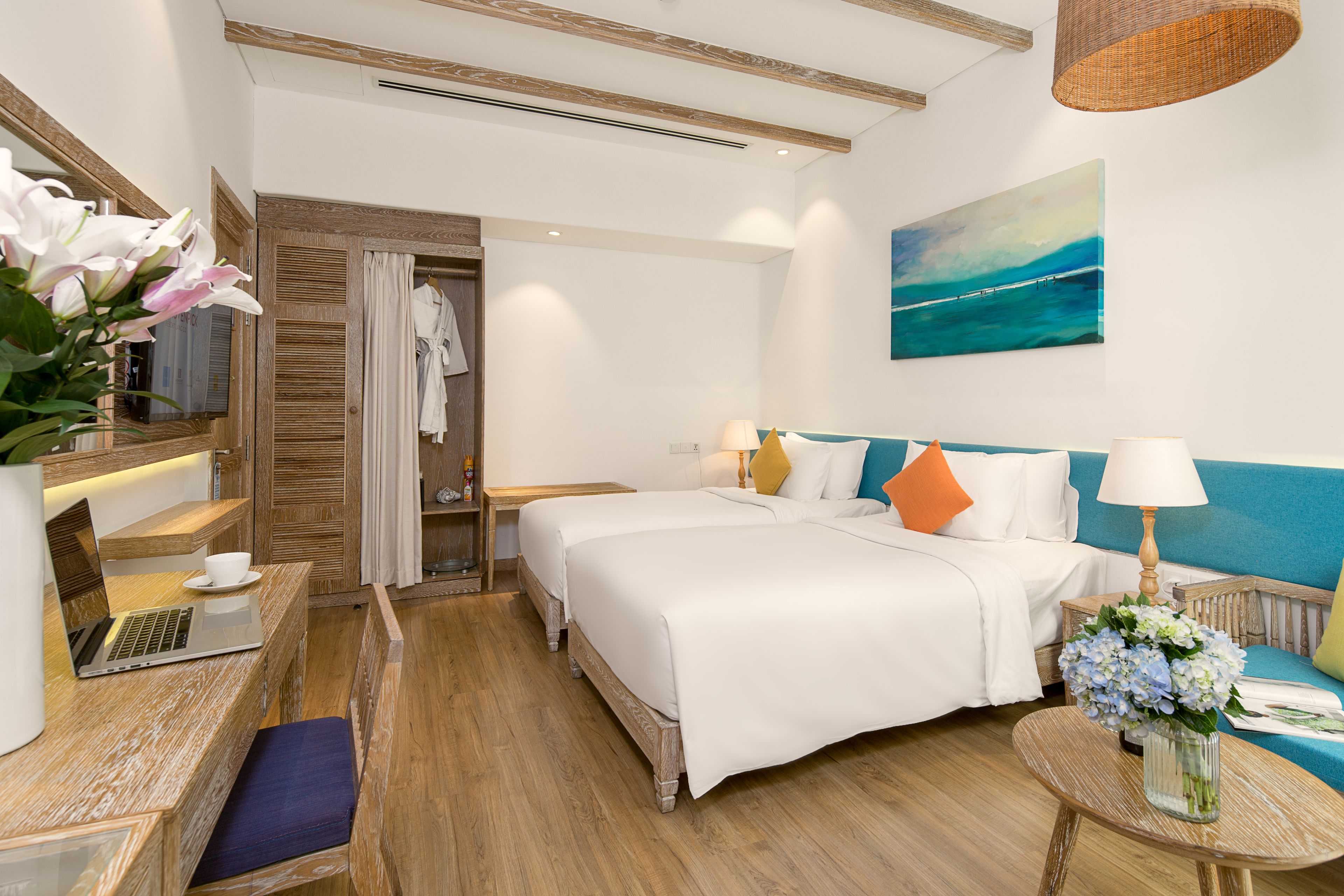 A hotel room with a large bed, a desk with a laptop, a seating area with a blue sofa, and a painting of a seascape on the wall.