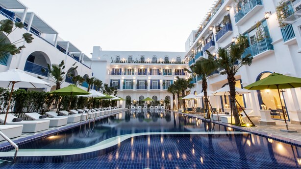 A hotel pool area with lounge chairs and umbrellas, surrounded by white buildings with blue balconies. - Risemount Premier Resort Da Nang (Da Nang)