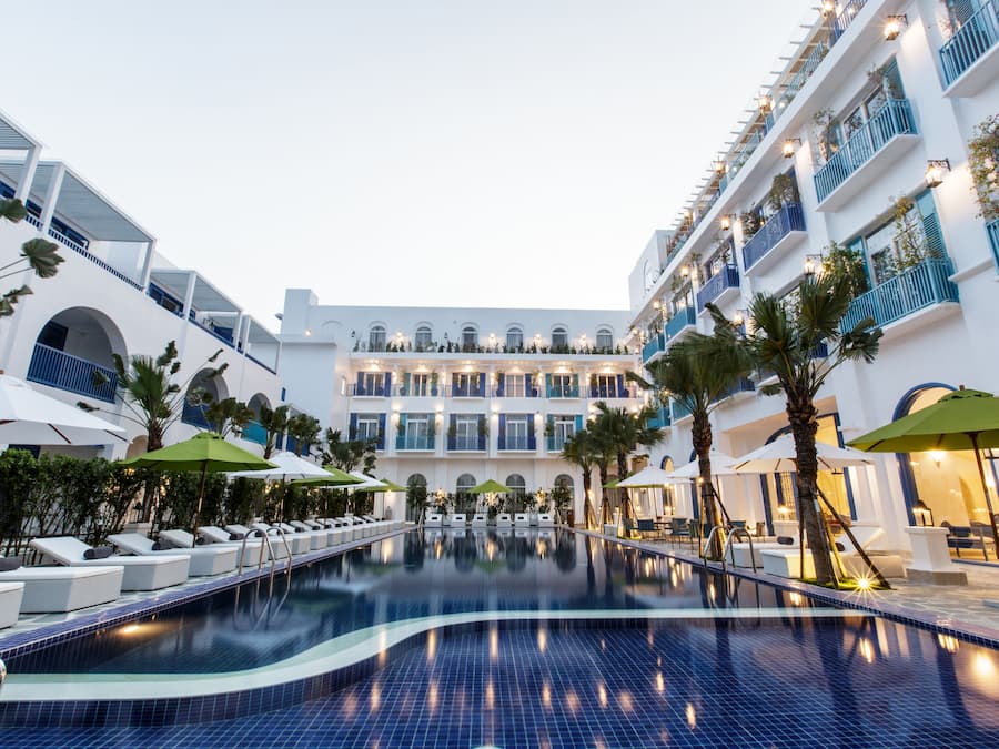 Zona de piscina de hotel con sillones y sombrillas, rodeada de edificios blancos con balcones azules.