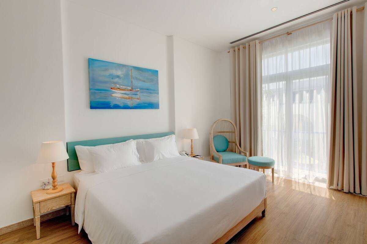 A neatly made bed with white linens, a painting of a sailboat on the wall, a wicker chair with a blue cushion, and a window with sheer curtains.